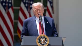 An older white man with blonde hair wearing a suit and speaking before a podium. US flags hang behind him.