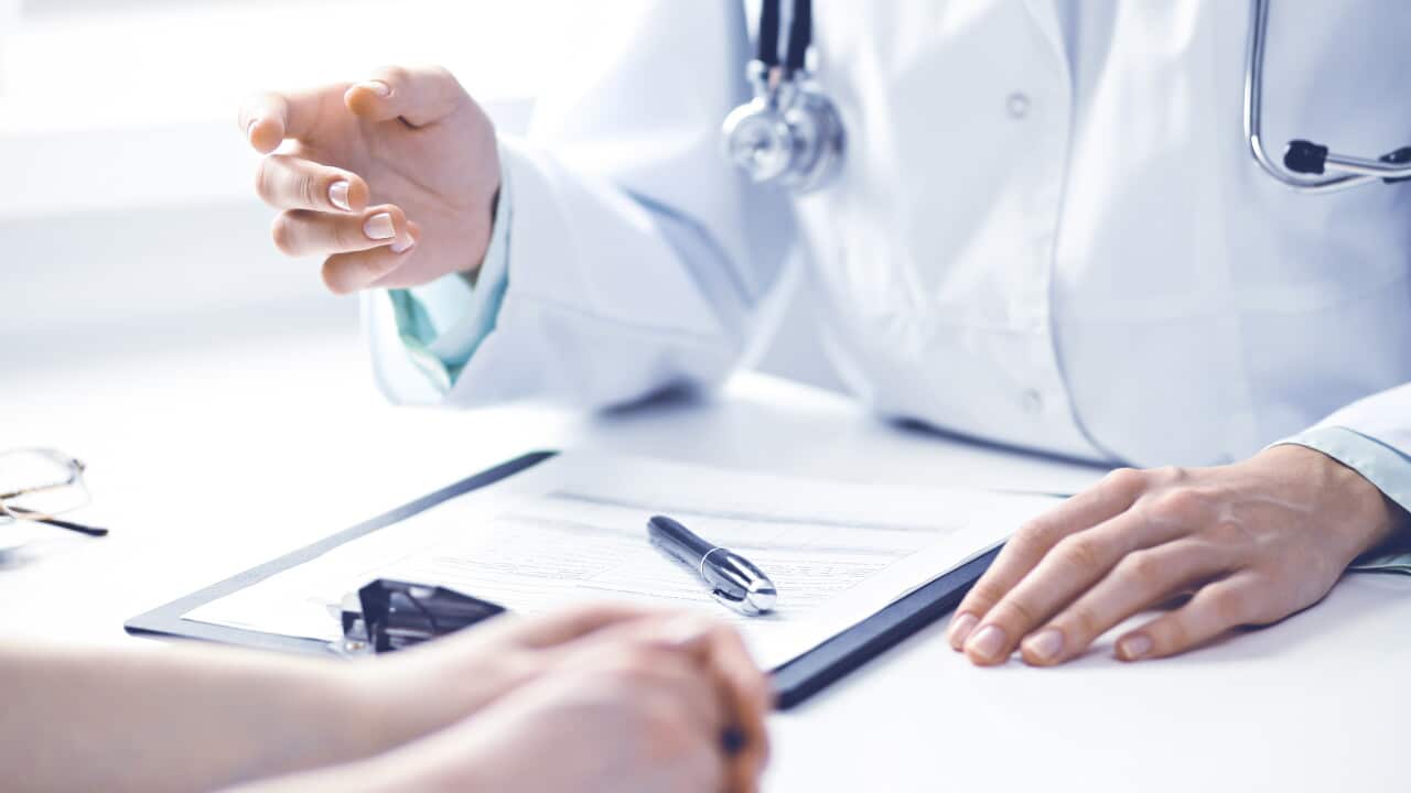 A close-up shot of a doctor in a white coat with a stethoscope around their neck, gesturing with their hand while speaking to a patient across a desk.