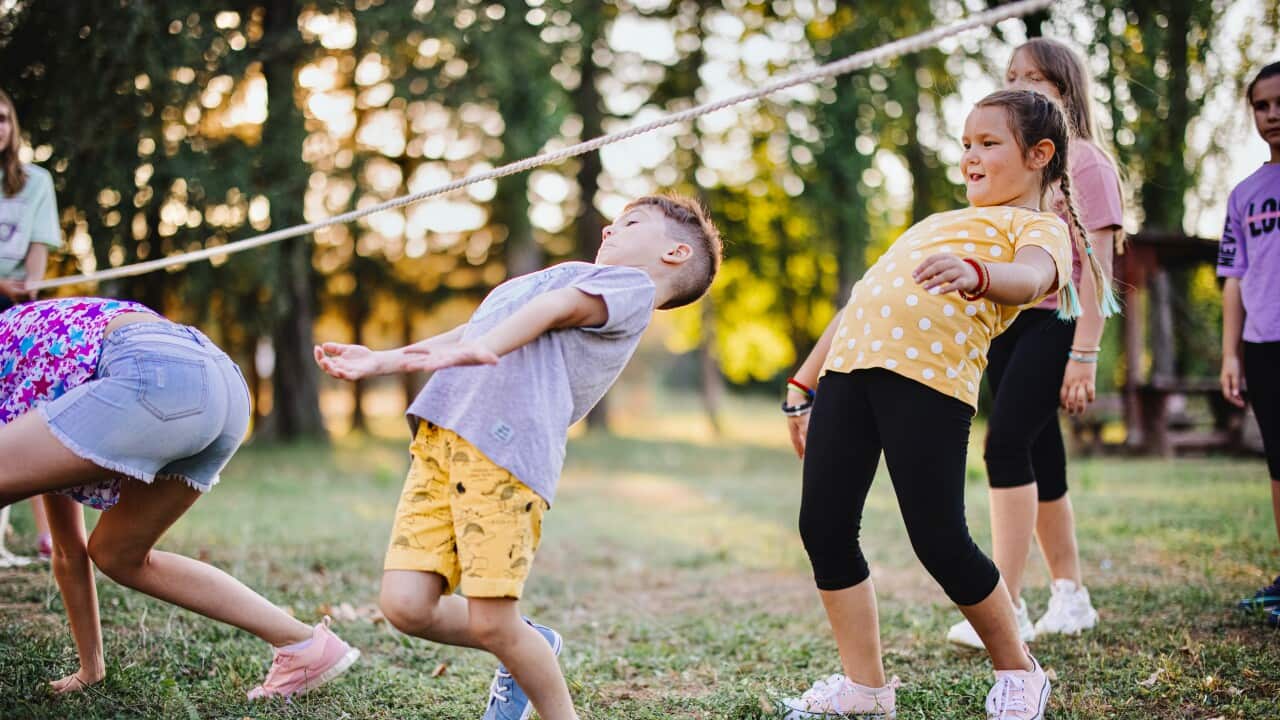Children in a line make their way under an outstretched rope, playing limbo.