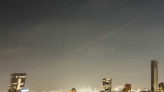 A wide night view of a city skyline lit up by glowing windows and streetlights, set beneath a hazy, dark sky that is cut diagonally by a single, thin orange light streak of light.