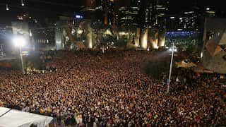 A massive crowd of people fills Federation Square in Melbourne at night, gathered in front of the illuminated, geometric facades of the buildings.