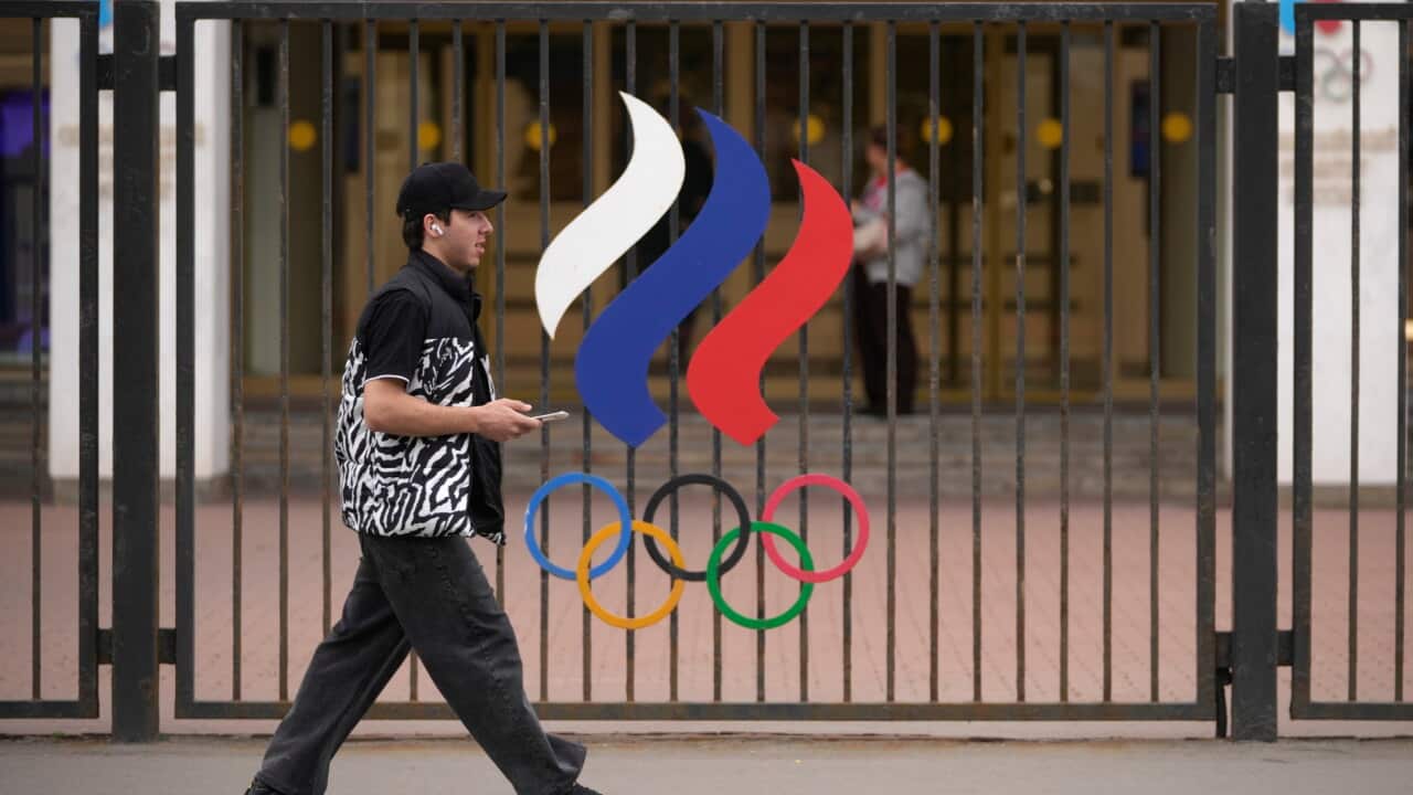 A pedestrian walks past an iron gate displaying the Olympic rings and a three-striped red-blue-and-white logo of the Russian Olympic Committee.