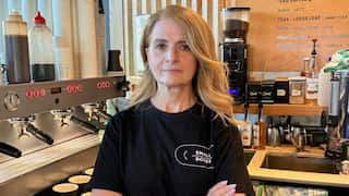 A woman with long blonde hair and wearng a black t-shirt stands with folded arms next to a coffee machine.