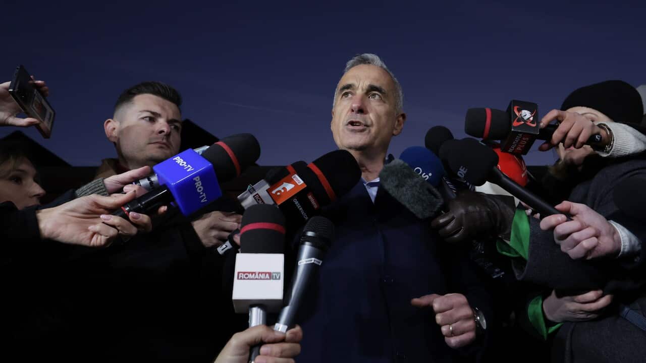 A man in a dark coat surrounded by reporters' microphones.