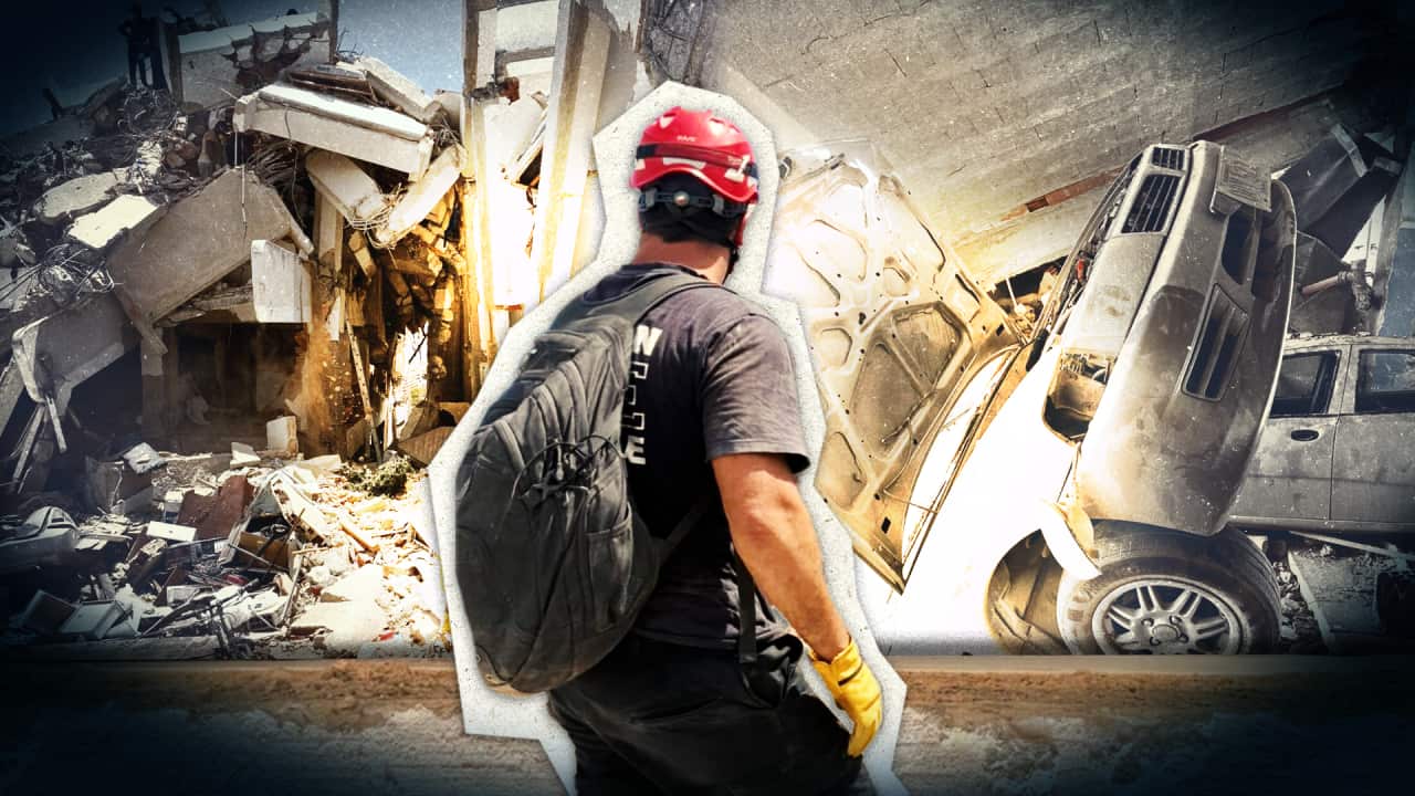 A stocky man in protective gear turns his back to the camera in front of disaster and destruction.