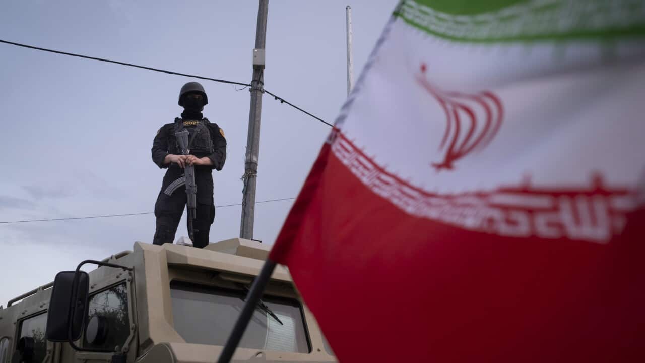 Masked armed officer standing on top of an armoured vehicle, with an Iranian flag blurred in the foreground.