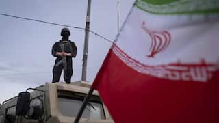 Masked armed officer standing on top of an armoured vehicle, with an Iranian flag blurred in the foreground.