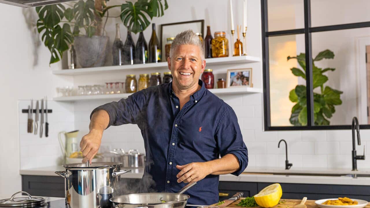 A smiling man stands in a kitchen, stirring something in a pot.