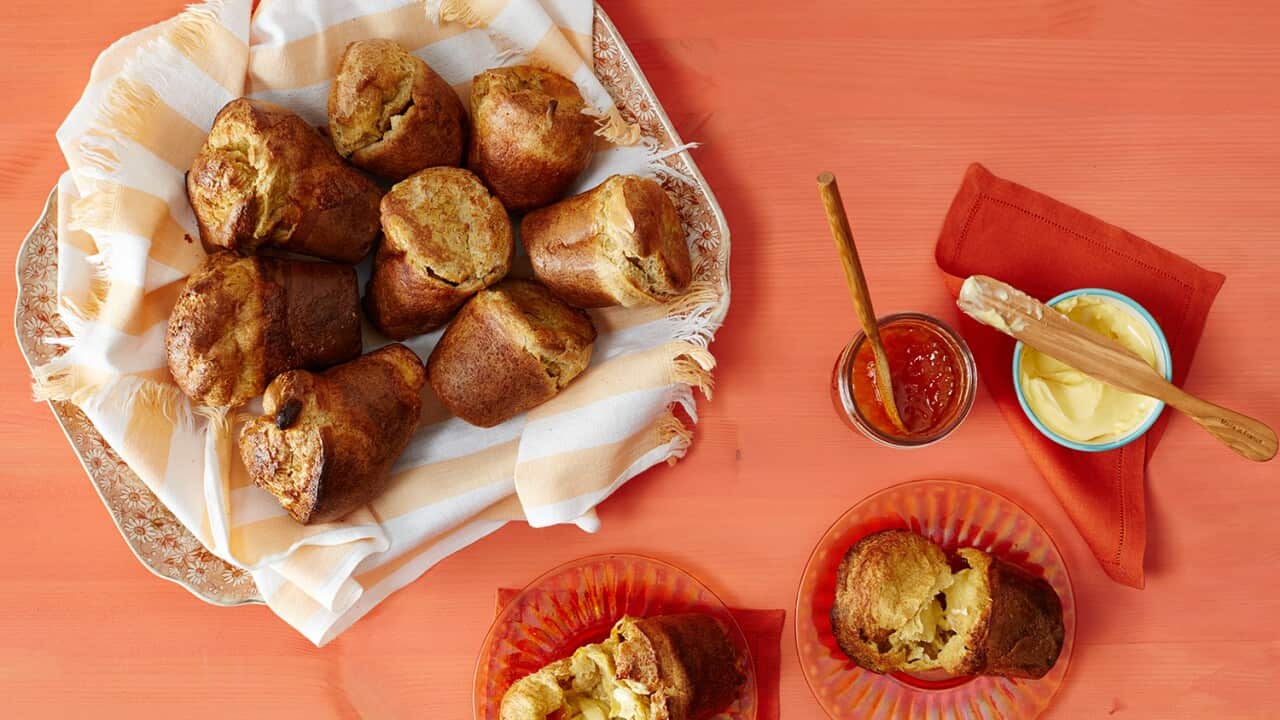 Seen from overhad, 8 golden-brown popovers, shaped like flower pots, sit on an apricot-and-white-striped napkin, on a floral plate. The plate,plus two torn-open popovers on small plates, a jam pot and some butter, all sit on an orange surface.