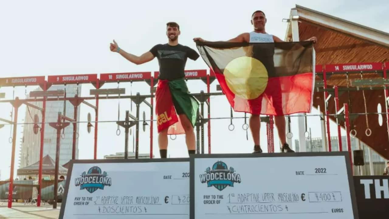 Eric Brown, who holds an aboriginal flag, and another man who has one arm stand atop the Wodcelona winning podiums amongst the outdoor gym equipment of the tournament.
