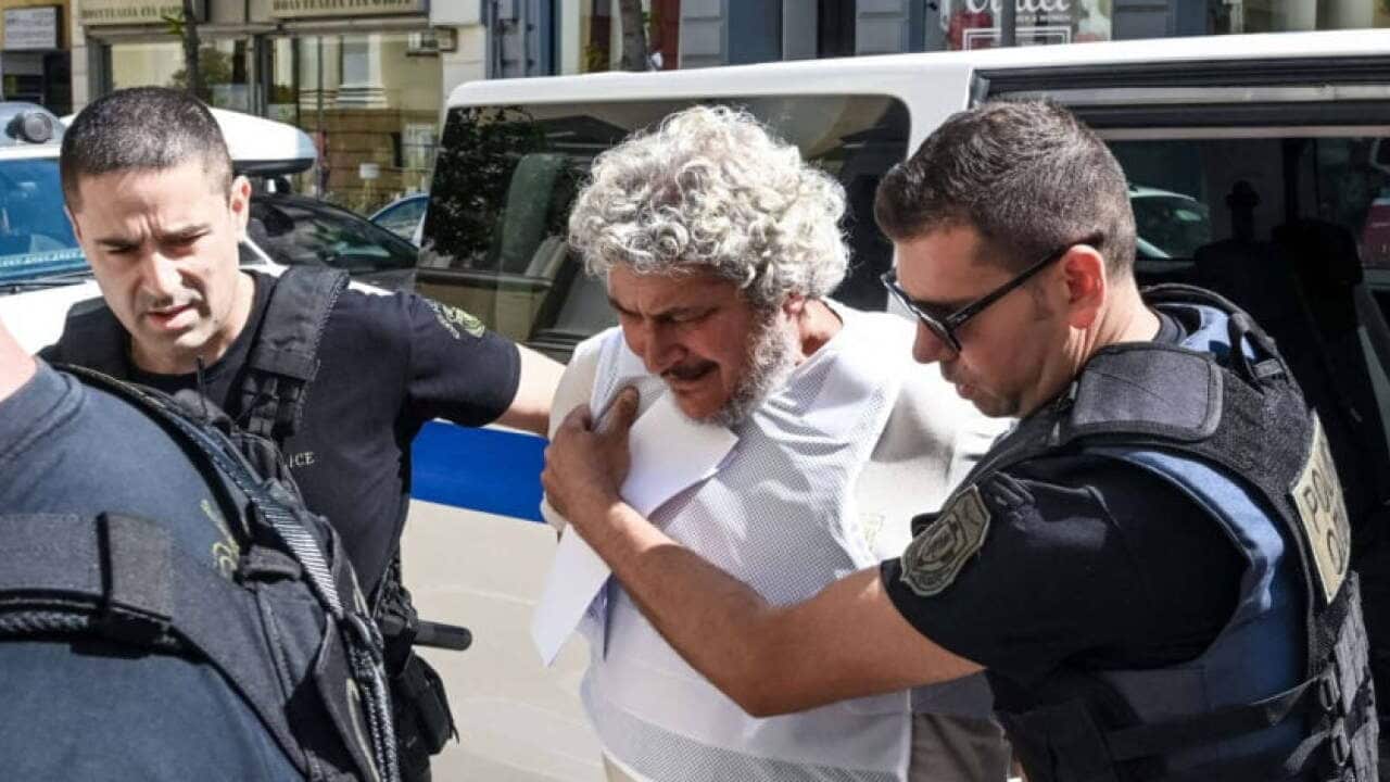 A man with curly grey hair and a beard is escorted by police officers on a city street.