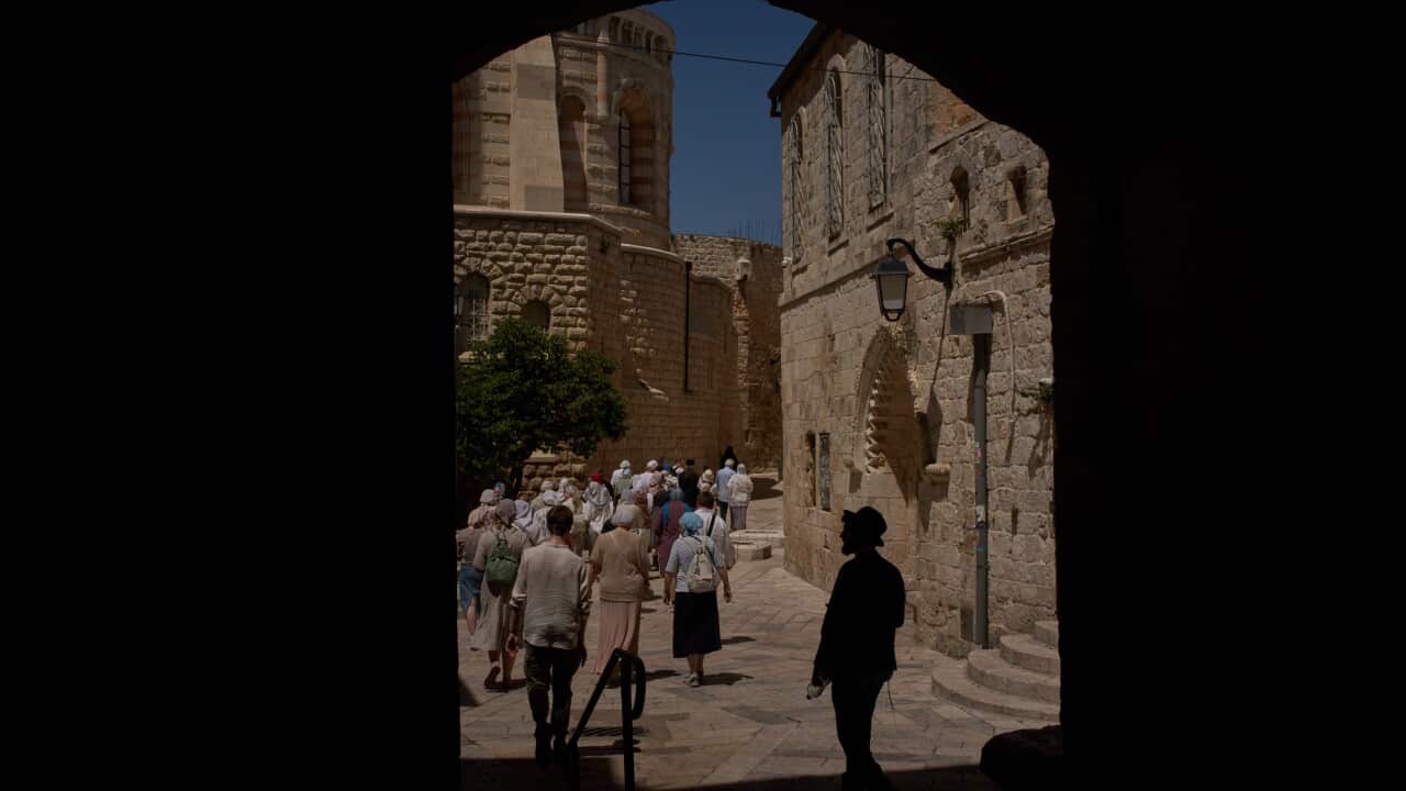 A group of tourists walk through a narrow stone street, seen from the shadow of a dark archway. Historic limestone buildings line the path, with bright sunlight illuminating the crowd and the old architecture beyond.