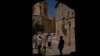 A group of tourists walk through a narrow stone street, seen from the shadow of a dark archway. Historic limestone buildings line the path, with bright sunlight illuminating the crowd and the old architecture beyond.