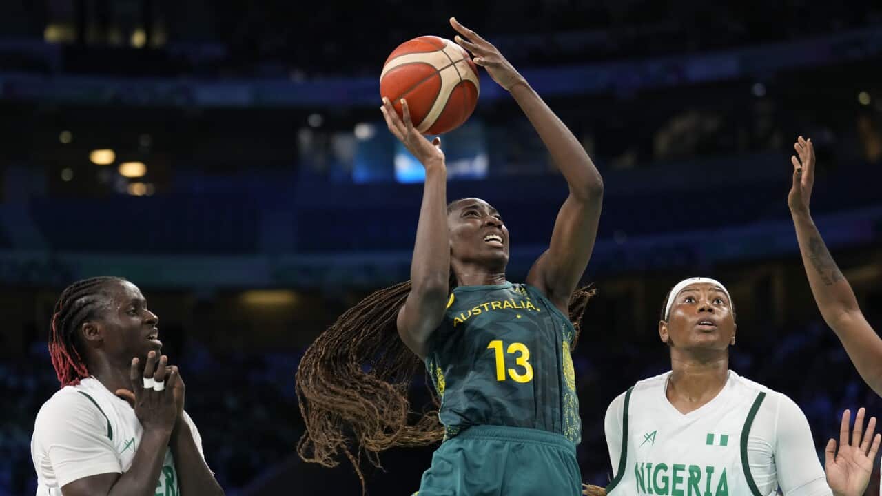 A female basketball player wearing a green uniform shooting as two players in white uniforms look on.
