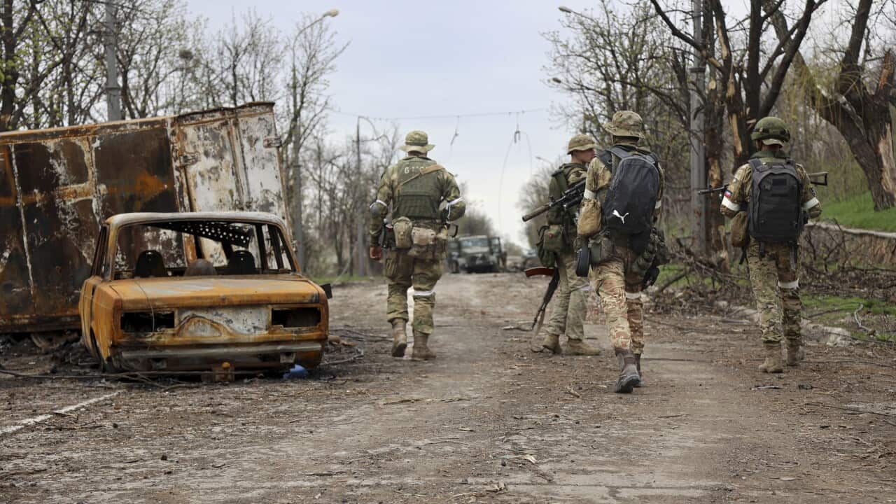 Four soldiers walk along a road amid destroyed vehicles.