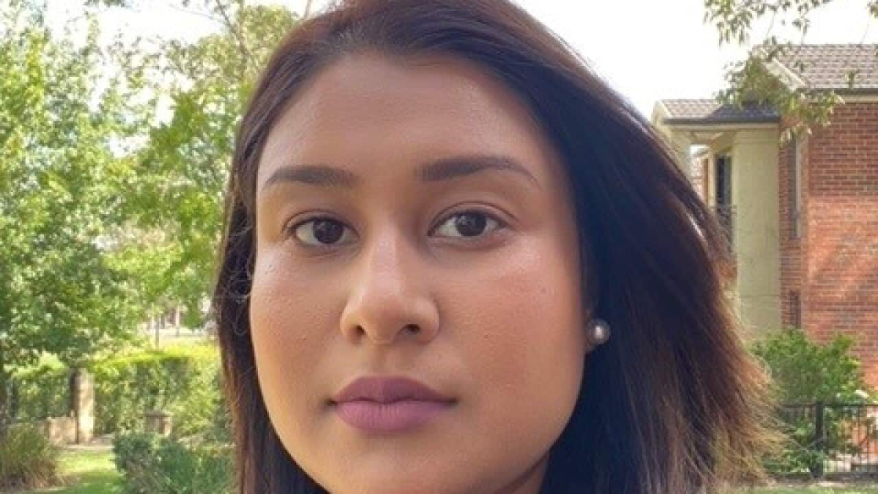 A close-up selfie of a woman with a neutral expression on a leafy street.