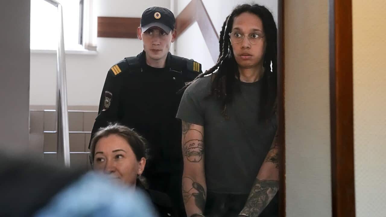 Woman dressed in prison clothing and handcuffs is escorted by a guard.