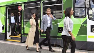 Prince Harry and Meghan Markle disembark a tram in Melbourne on a royal visit to Australia. Harry, a tall man with red hair and beard, is dressed in a grey blazer with black slacks. Megan, a woman with dark hair, is wearing a black dress with a tan coloured trenchcoat and ballet flats.