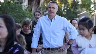 A man in a light blue button-down shirt stands outdoors among a small crowd, resting one hand on a stroller and the other on his hip.