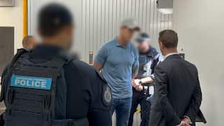 An Australian Federal Police officer stands in the foreground, facing several men and an official from the Office of the Special Investigator in an indoor corridor.