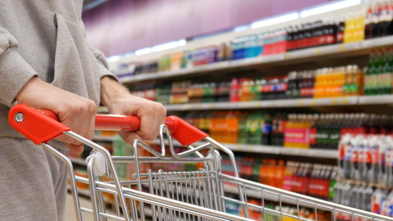 A person holding a shopping trolley in front of a row of sugary drinks at a supermarket.