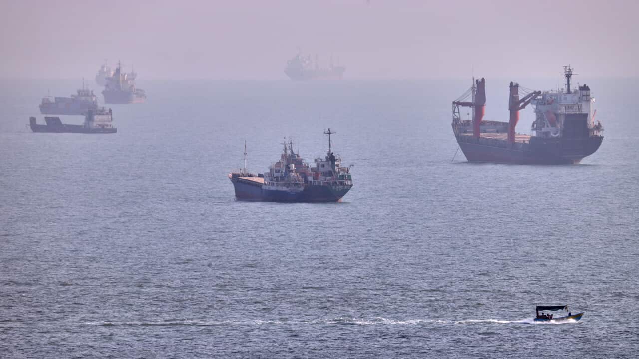 Several cargo ships and smaller vessels sit on a hazy sea, while a small motorboat travels across the foreground, leaving a white wake behind it.