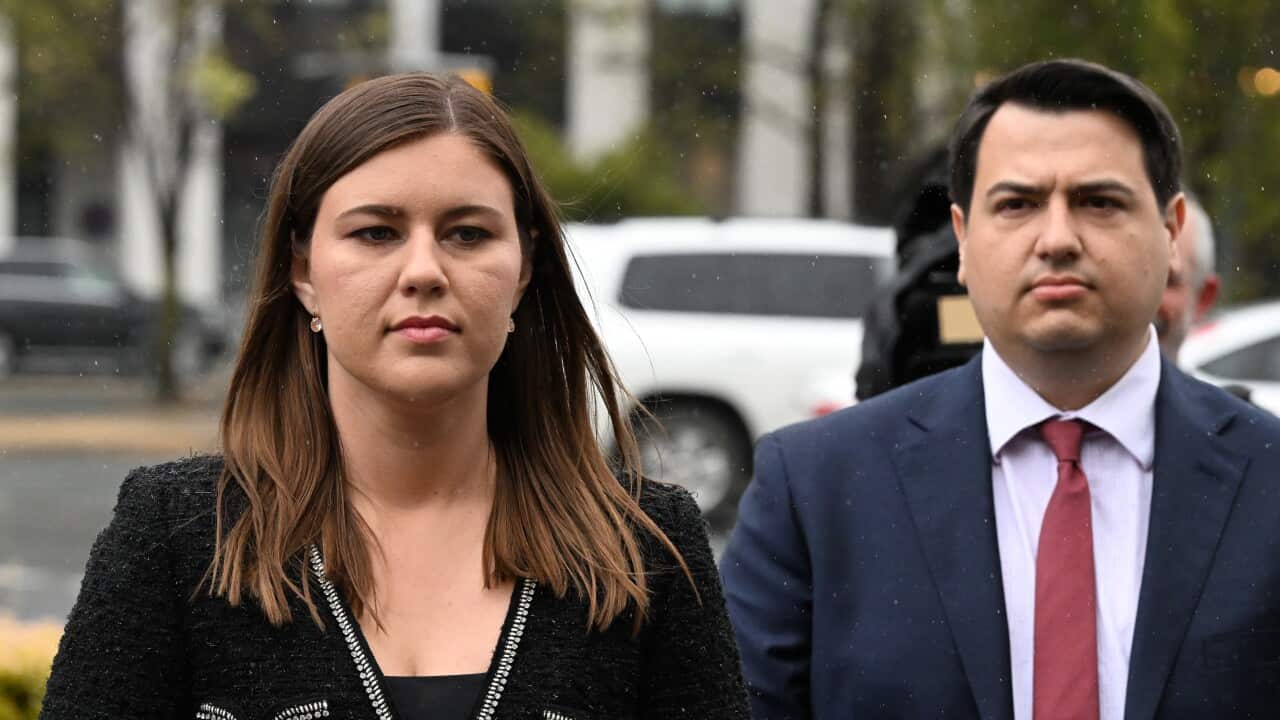 A man in a suit and red tie walks behind a woman in a black suit.