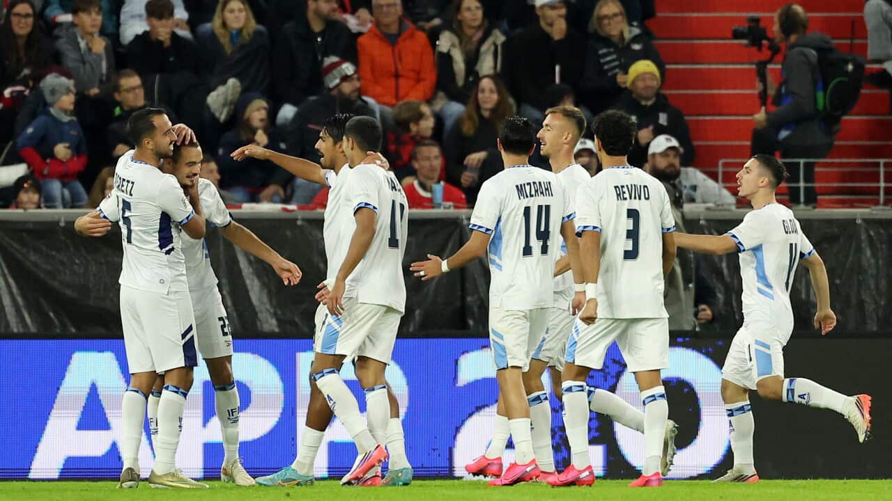 Football players in white uniforms celebrate on the field during a match, with teammates gathering together as spectators watch from the stadium stands behind them.