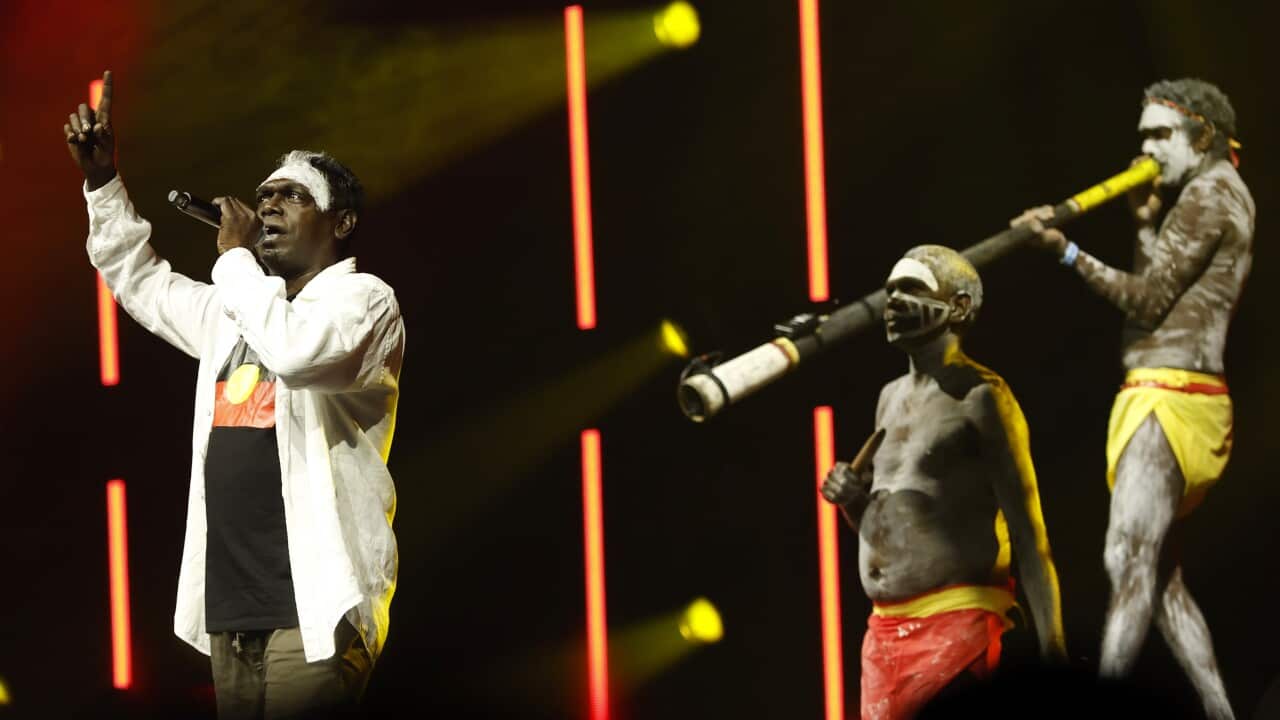 An Aboriginal Australian singer in a white long-sleeved collared shirt points his finger upwards on a dark stage with red and yellow lights, while three men in body paint and red and yellow loincloths perform beside him, one playing a didgeridoo.