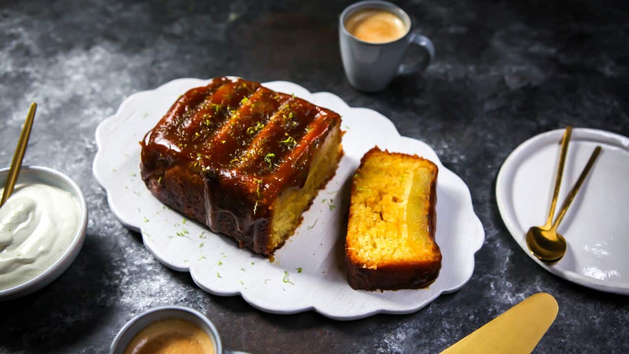 A loaf cake sits on a scallop-edged white platter. A slice has been cut off, showing a sticky top layer. The whoel cake is coated with a sticky glaze. Cream sits in a bowl nearby.