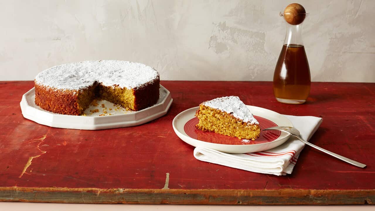 A round, low yellow cake dusted with icing sugar sits on a white plate on a red board. A slice of cake on a red and white plate also sits on the board, with a fork propped on the plate edge and a napkin underneath.
