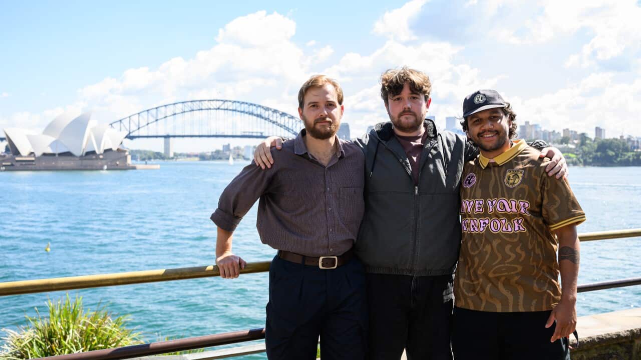 Three men stand together with their arms around each other, posing for a photo along a waterfront railing. In the background, the Sydney Opera House and the Sydney Harbour Bridge are visible under a bright, partly cloudy sky. The man on the left wears a brown button-down shirt and navy trousers, the man in the center wears a grey zip-up jacket, and the man on the right wears a patterned brown shirt with the words "LOVE YOUR KINFOLK" and a black baseball cap.