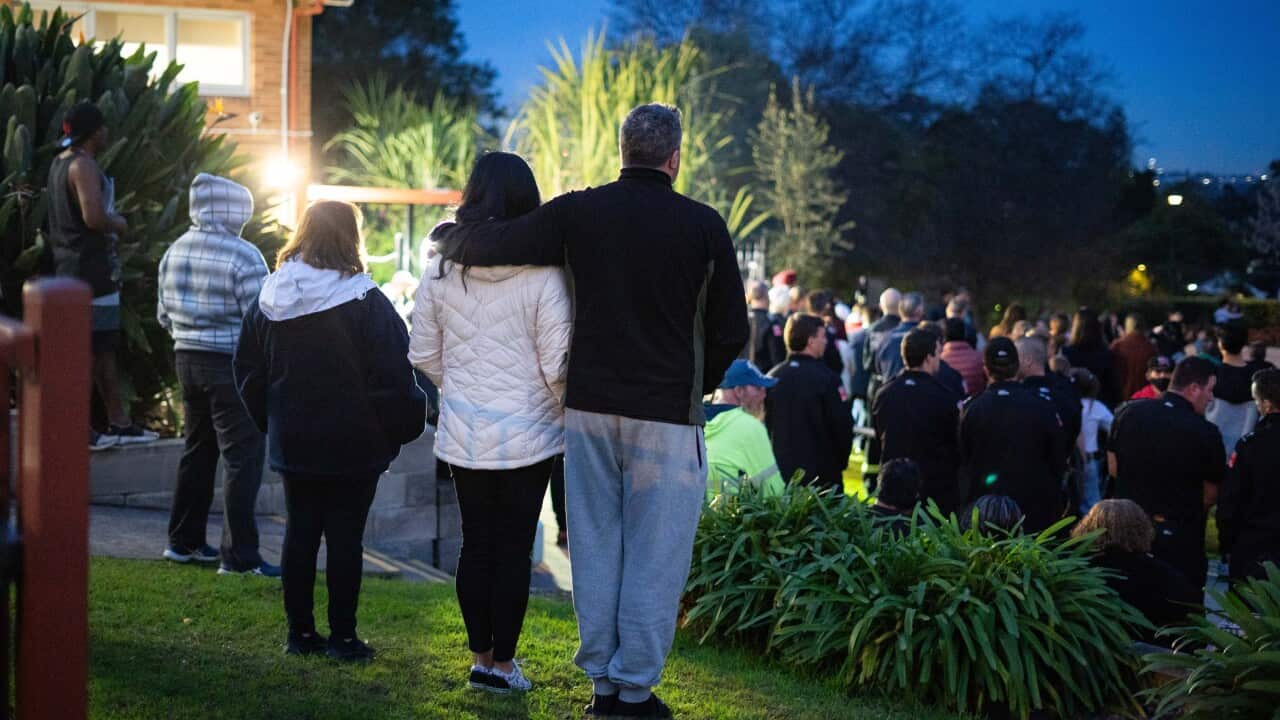 In the foreground, a man stands with his arm around a woman as night falls over a gathered crowd in the background.