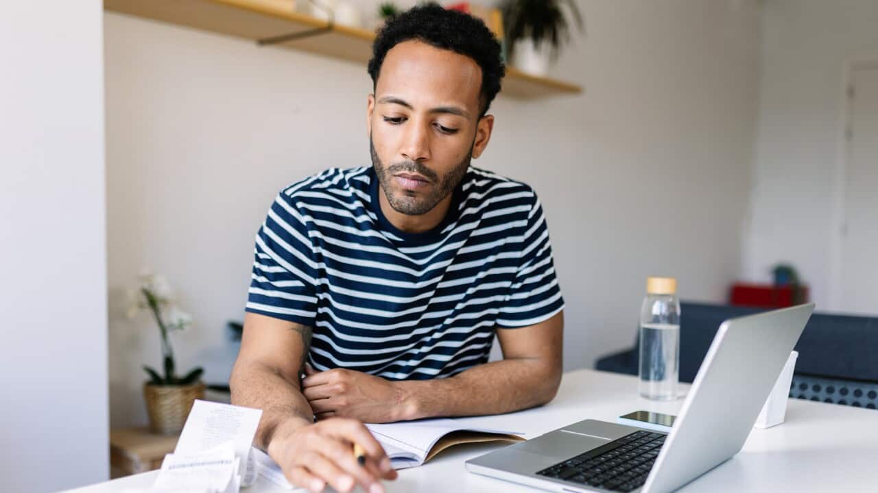 Young adult man with laptop checking bills, taxes, bank account balance and calculating expenses sitting at living room table.