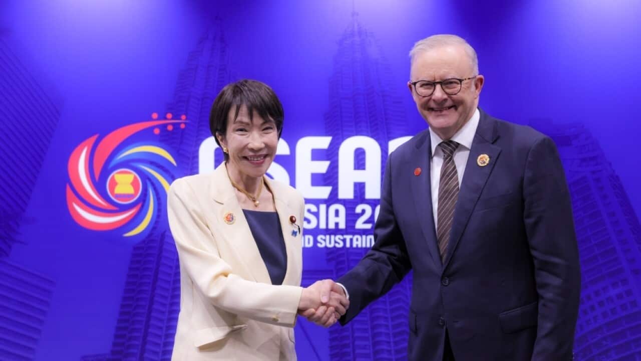 A man and a woman shaking hands, smiling at the camera in front of a blue wall
