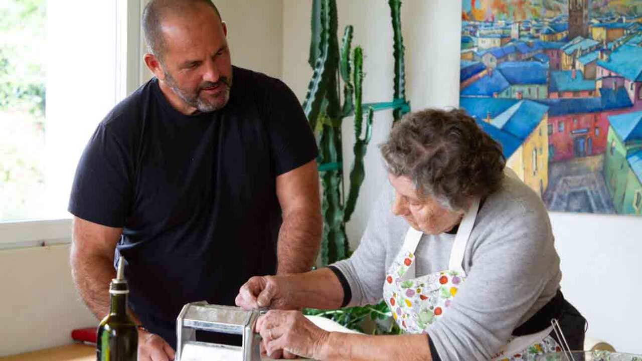 A man in a black t-shirt stands in a home kitchen beside an older woman in an apron, who is working a pasta rolling machine.