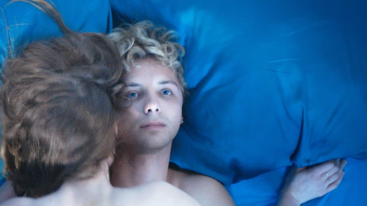A person with short curly hair lies on a blue sheet, staring at the camera, with another person lying over them.