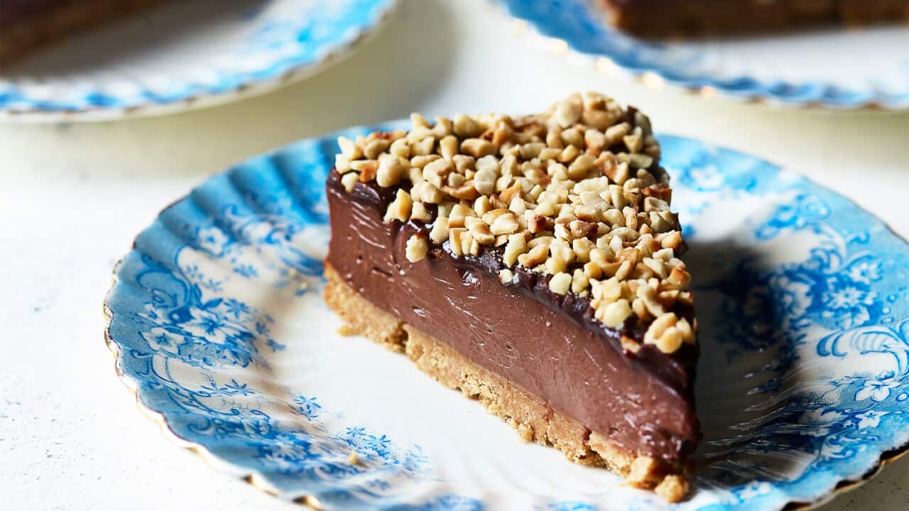 A wedge-shaped slice of cheesecake sits on a pretty plate with a textured and blue-patterned edge. The cheesecake is rich and lush, with a biscuit base, a deep middle layer and a thin darker top layer, sprinkled with chopped nuts. Two silver forks sit in front of the plate. Two more plates, with slices of cheesecakes, can be seen in the background.