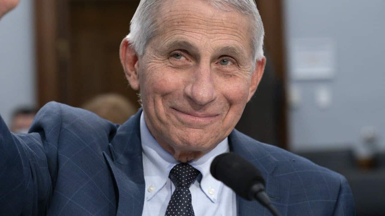 Anthony Fauci sitting at a desk in front of a microphone and raising his hand.