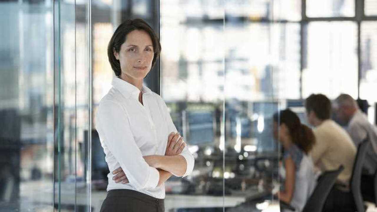 Businesswoman standing in office with group of office workers in background.