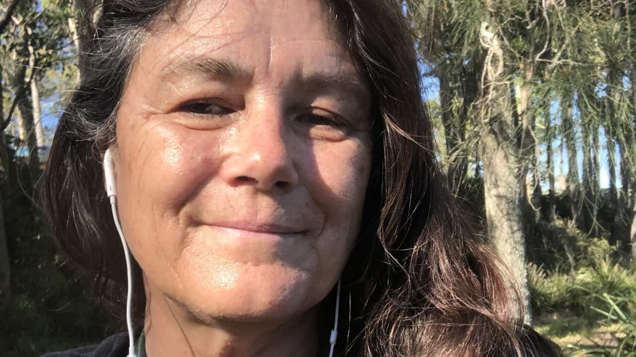 A middle-aged woman wearing a black t-shirt and with headphones in her ears outside with trees in the background