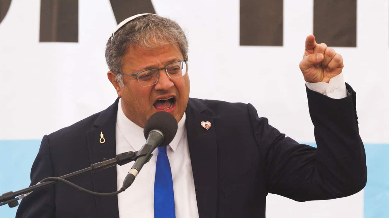 A man with glasses, grey-streaked hair, and a white kippah wears a dark suit with a blue tie as he speaks passionately into a microphone with his left hand raised making a pointing gesture.
