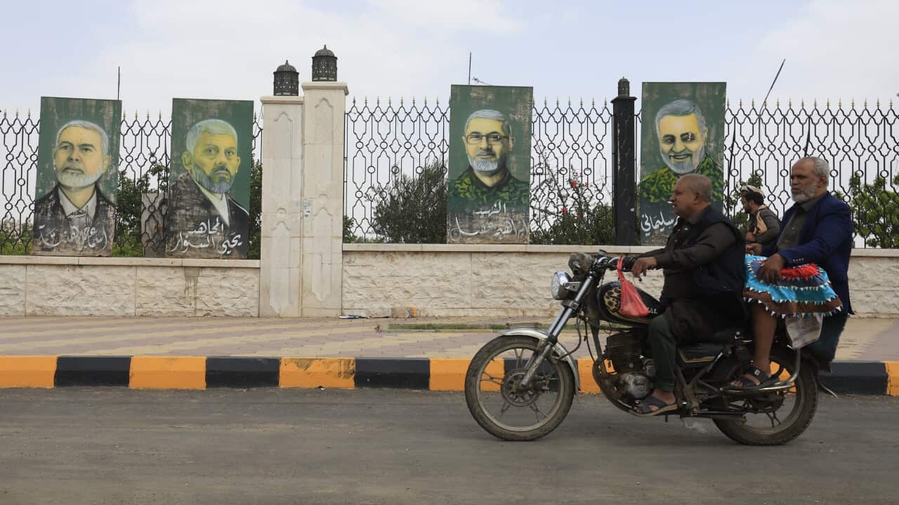 Two men ride a motorcycle past a fence displaying painted portraits on tall panels, with a paved street and decorative metal fencing in the background.