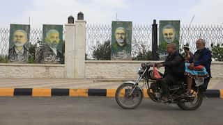 Two men ride a motorcycle past a fence displaying painted portraits on tall panels, with a paved street and decorative metal fencing in the background.