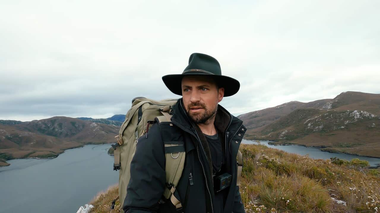 A man in a wide hat, wearing a jacket and backpack, stands in front of a sweeping vista, with water and low mountains behind him.