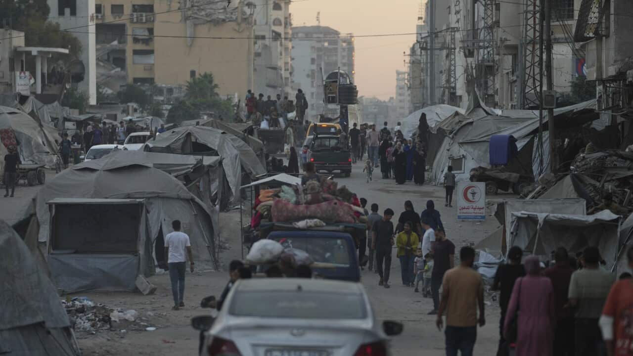 Cars and displaced people walk along a street, past tents.