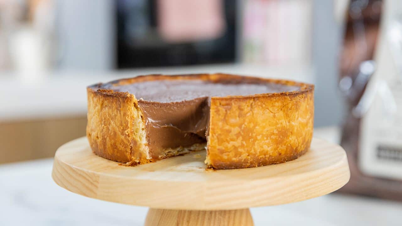 A deep tart with a dark golden-brown pastry case sits on a light-coloured wooden cake stand. A piece has been cut and removed from the flan, showing the baked chocolate custard filling.