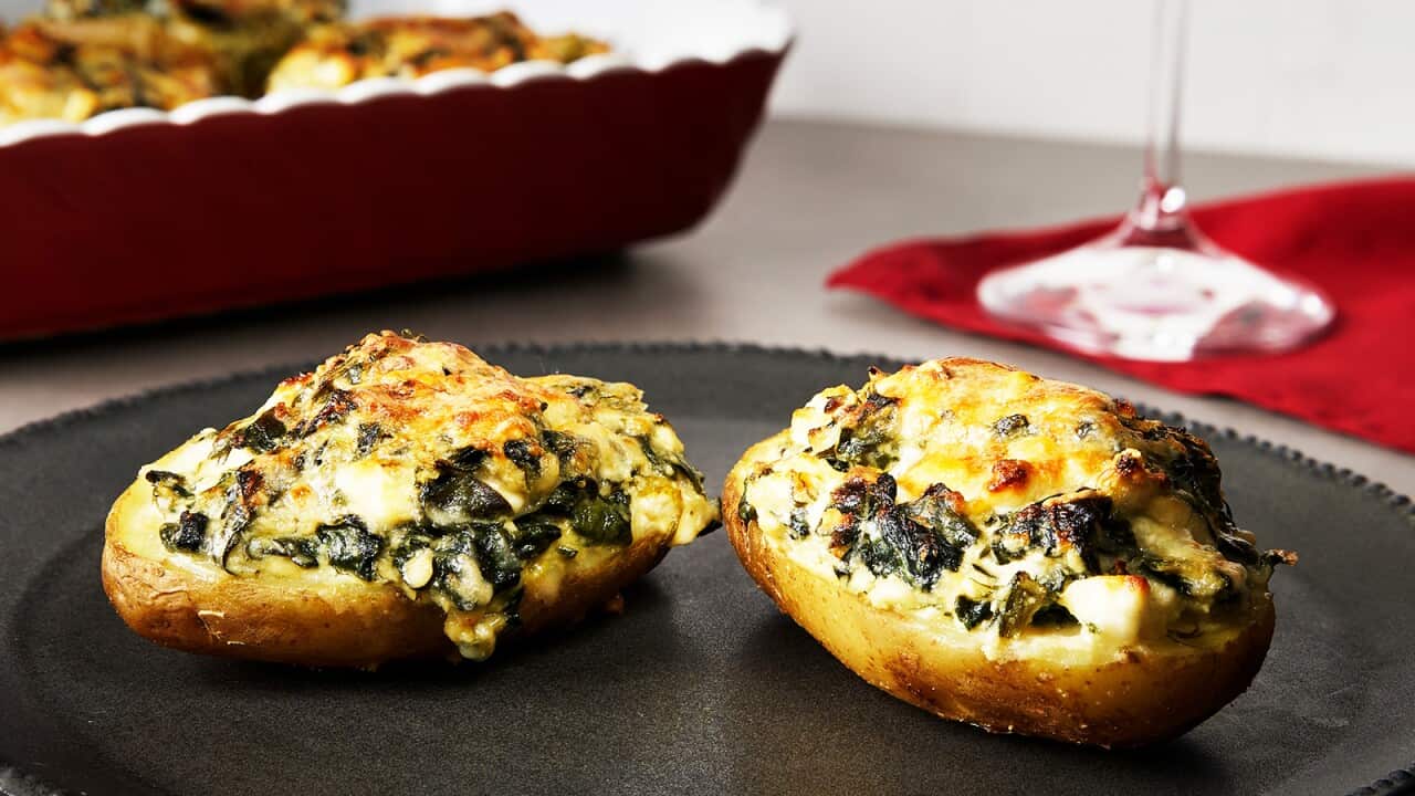 Two potato halves sit on a round dark metal tray with a narrow decorative edge. Each potato half is topped with a spinach and cheese mixture, with the cheese gone golden brown on the top. A baking dish with more potato halves sits in the background.
