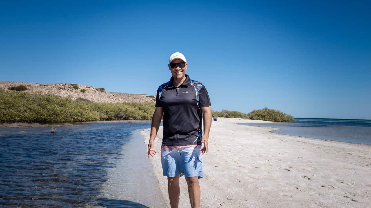 Michael Wear, a Malgana man in his 40s wearing a black t-shirt, pale blue shorts and a white cap, stands on a shite sandy beach flanked by ocean.