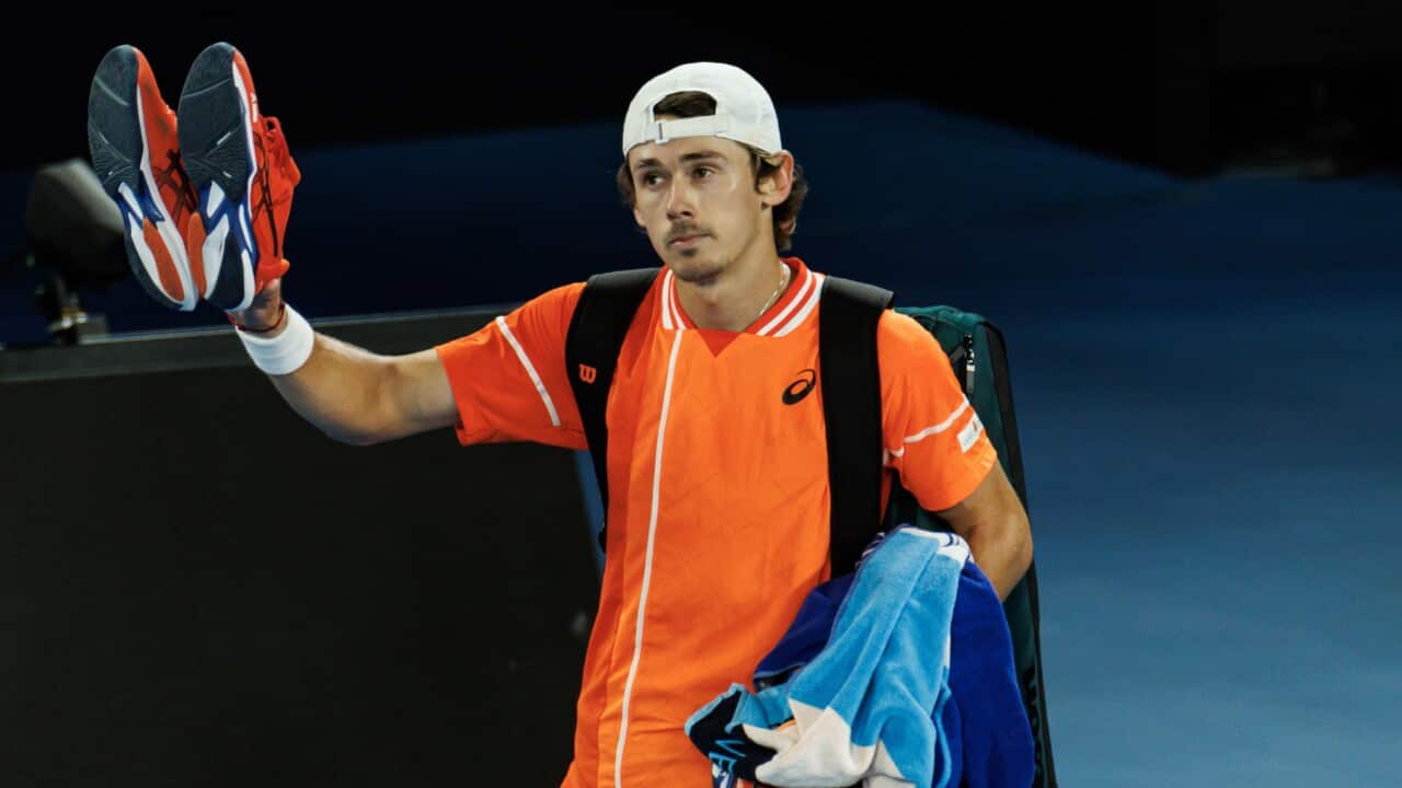 Alex de Minaur holds his shoes up in a wave to viewers after a match. He is dressed in orange.